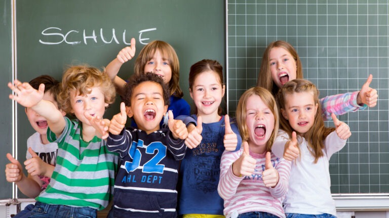 Eine Gruppe von Grundschülern, die freudig vor einer Schultafel stehen, auf welcher das Wort 'Schule' geschrieben steht.