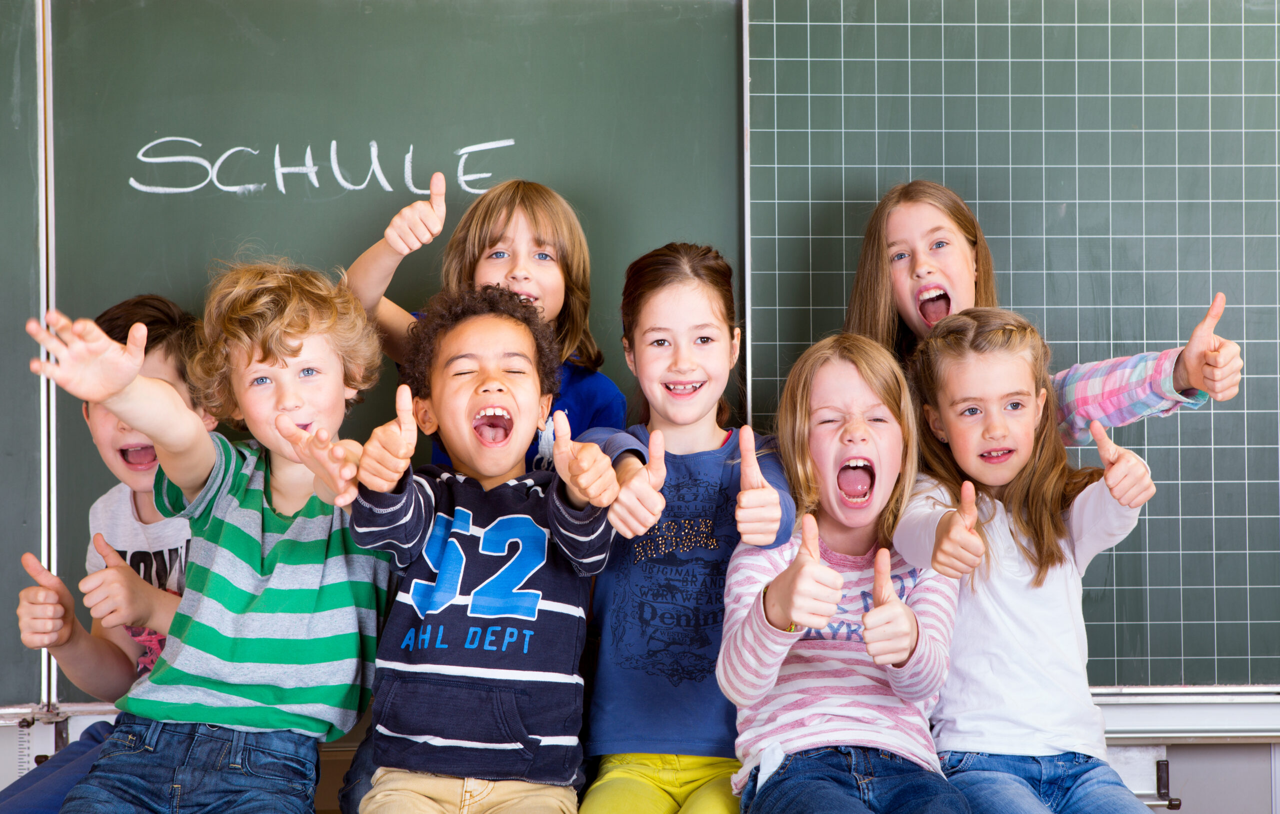 Eine Gruppe von Grundschülern, die freudig vor einer Schultafel stehen, auf welcher das Wort 'Schule' geschrieben steht.